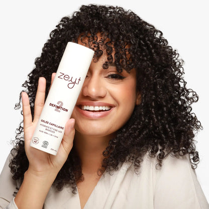 Femme souriante avec des cheveux bouclés, tenant une bouteille de gelée capillaire "Definition 2" de Zeyt. Cette gelée hydrate et fixe les boucles avec des ingrédients naturels comme l'aloe vera, le lin et le chia.