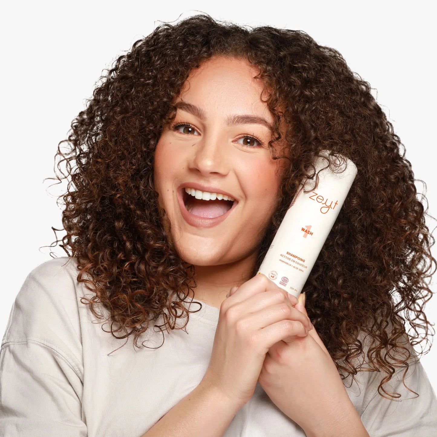 Femme souriante aux cheveux bouclés, tenant une bouteille de shampoing "Wash 1” de Zeyt. Ce shampoing nettoie en douceur les cheveux avec des ingrédients naturels comme l'hamamélis et l'aloe vera.